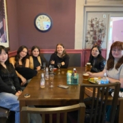 Six people are seated at a table indoors, smiling at the camera, with drinks and a casual dining setup, in a room with purple walls and artwork.