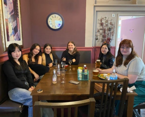 Six people are seated at a table indoors, smiling at the camera, with drinks and a casual dining setup, in a room with purple walls and artwork.