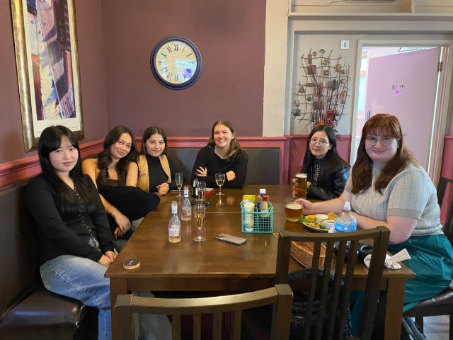 Six people are seated at a table indoors, smiling at the camera, with drinks and a casual dining setup, in a room with purple walls and artwork.