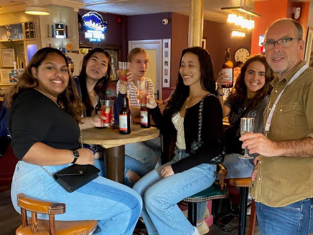 A group of six people enjoying drinks at a pub, smiling at the camera, with a person on the right holding a glass.