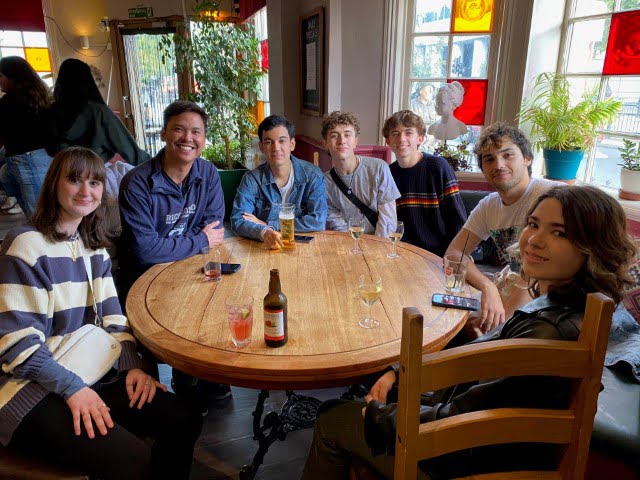 A group of seven people is sitting around a wooden table with drinks in a warmly lit room with plants and colored glass windows.