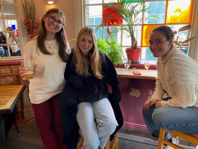 Three people are posing inside a cozy, well-lit room with a wooden bar, plants by the window, and colorful stained glass panels.