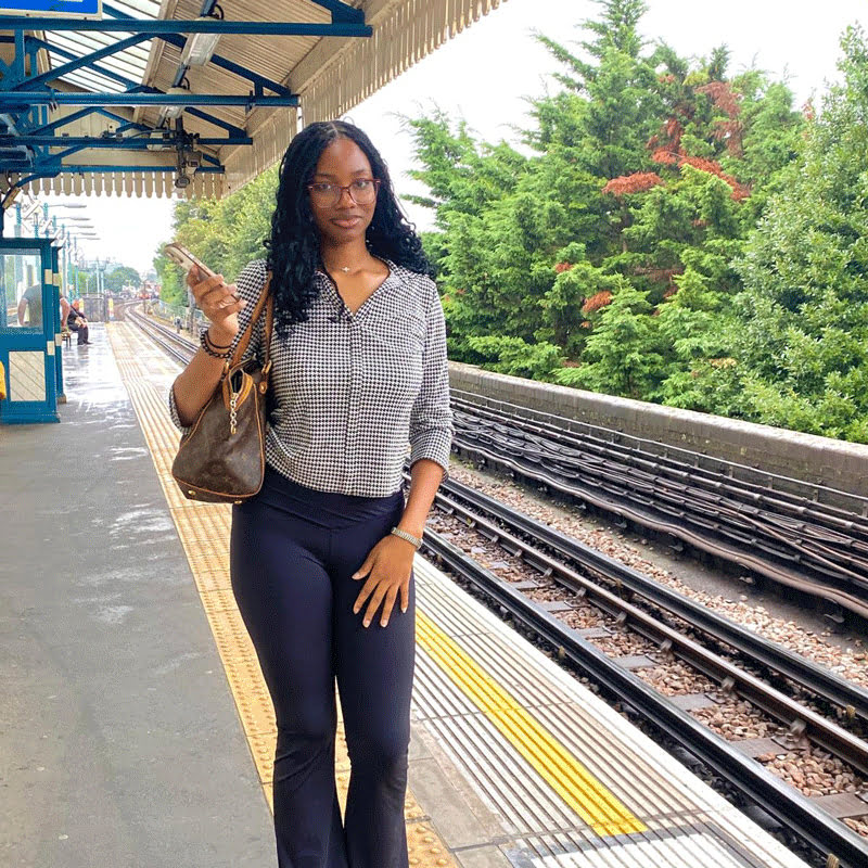 A person stands on a train platform holding a phone, surrounded by greenery. The platform appears empty aside from distant figures.