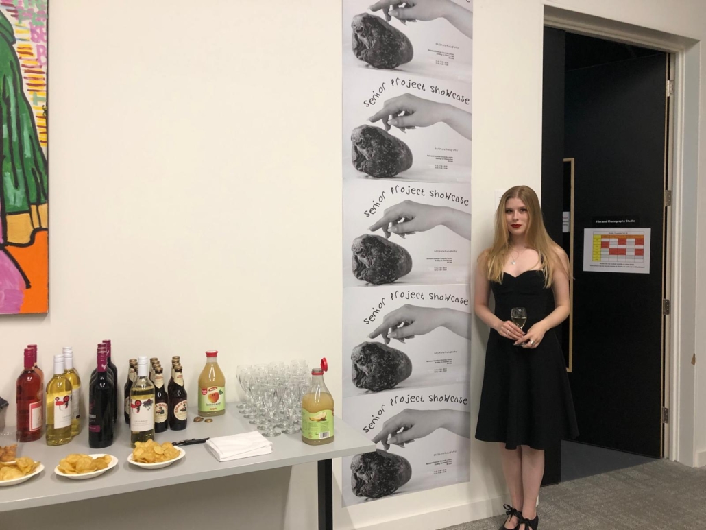 A person in black dress holds a glass near a table with beverages and snacks, next to "Senior Project Showcase" posters.