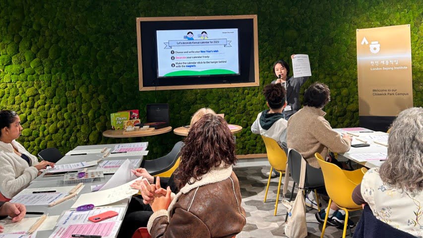 A person leads a workshop at London Beijing Institute, Chiswick Park Campus. Participants engage with materials on a table, surrounded by a moss wall.