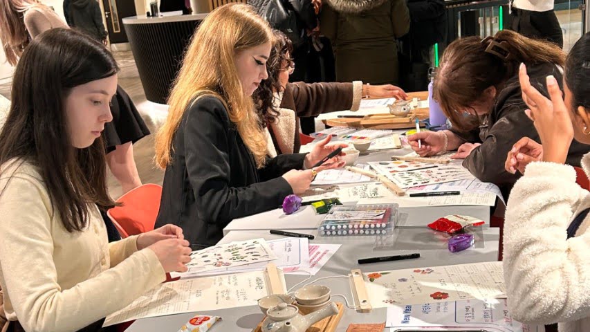 People engaged in arts and crafts at a table, working on papers with colorful materials. The setting suggests a creative, collaborative environment.