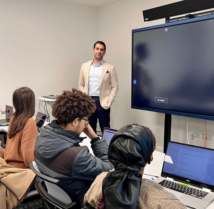 A person stands near a screen displaying "No signal" while three others seated use laptops in a classroom setting.
