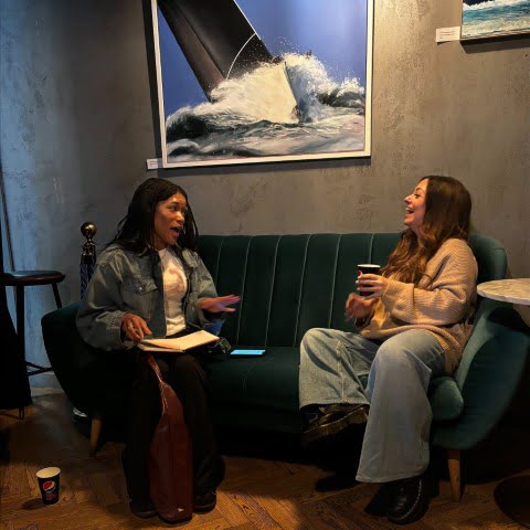 Two people sit on a couch in a modern café, conversing. A painting of ocean waves hangs on the wall behind them.