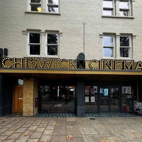 The image shows the entrance of Chiswick Cinema, featuring a marquee with illuminated lettering. It is located in an urban setting.