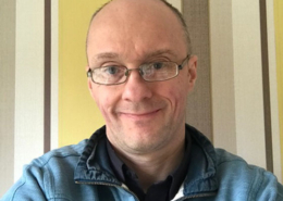 A person wearing glasses and a blue jacket stands in front of a striped wall, smiling at the camera.
