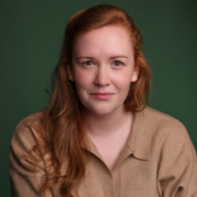 A person with long red hair and a beige shirt stands against a green backdrop, looking directly at the camera with a slight smile.