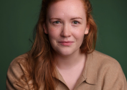 A person with long red hair and a beige shirt stands against a green backdrop, looking directly at the camera with a slight smile.
