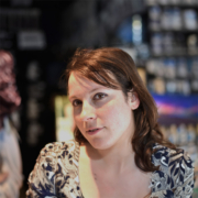 A person with brown hair stands in a dimly lit room, surrounded by blurred shelves, wearing a patterned shirt.