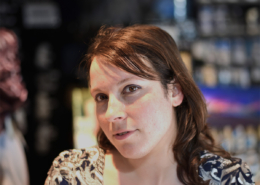 A person with brown hair stands in a dimly lit room, surrounded by blurred shelves, wearing a patterned shirt.