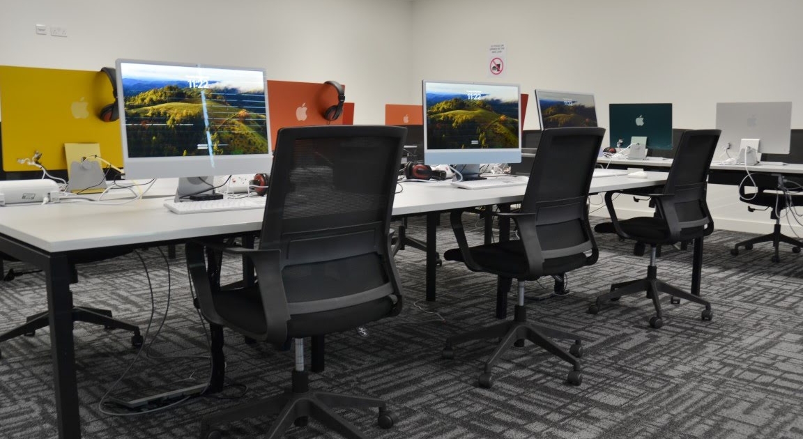 A modern computer lab with rows of Apple desktops on tables, ergonomic chairs, and a patterned carpet in a well-lit room.