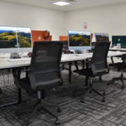 A modern computer lab with rows of Apple desktops on tables, ergonomic chairs, and a patterned carpet in a well-lit room.
