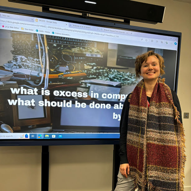 A person stands smiling in front of a digital presentation discussing computing excess. A large screen displays the topic with colorful imagery.