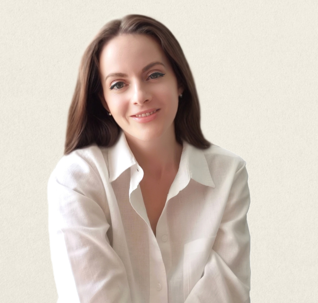 A person with long hair, smiles at the camera, wearing a professional white shirt. The background is plain and neutral-toned.