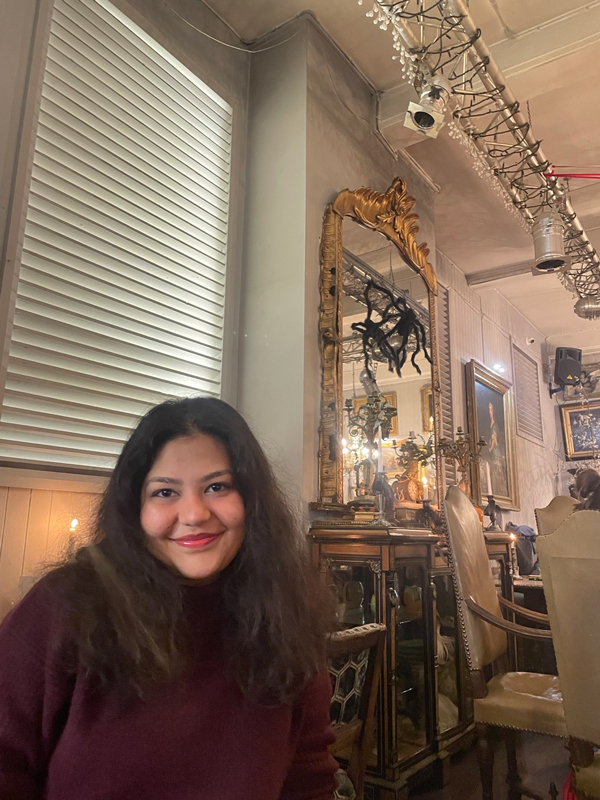 A person with dark hair smiles in an ornate, vintage-style room with mirrors, paintings, and elegant furniture. The ambiance is cozy and inviting.