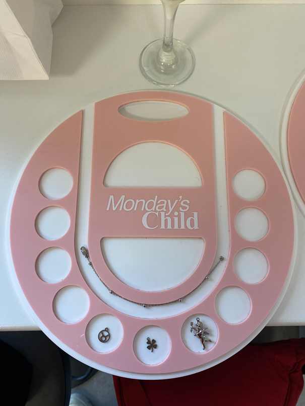 A pink beading board with "Monday's Child" text, various small charms, and a wine glass in the background on a white surface.