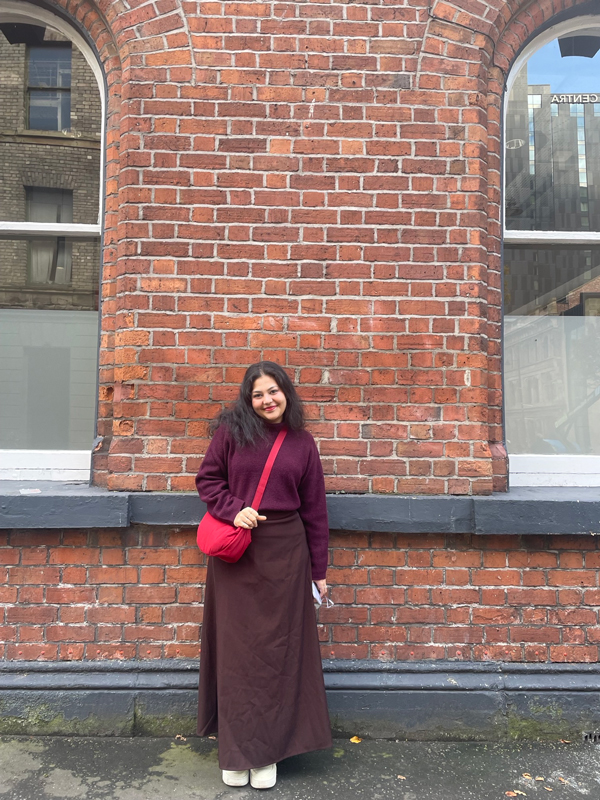 A person stands smiling against a red brick wall with large windows, holding a red bag. The scene is urban and casual.