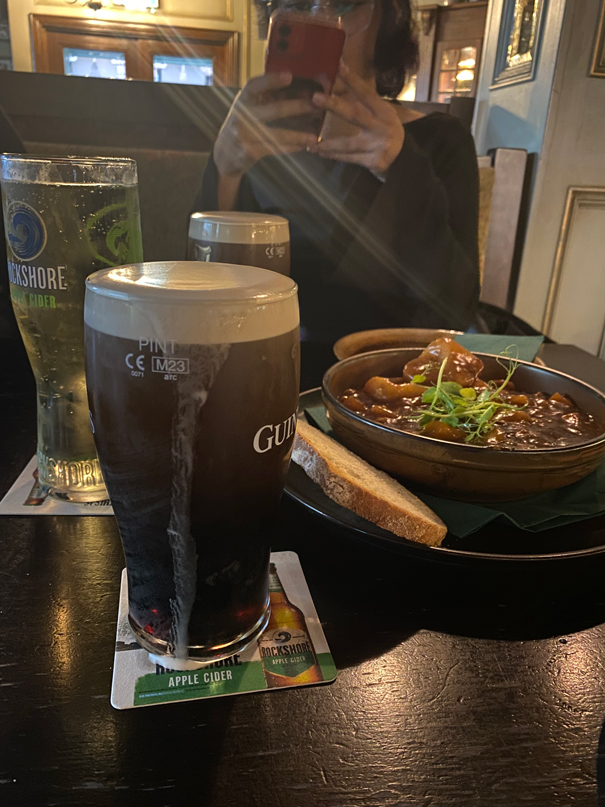 A person holds a phone in a pub, with pints of beer, a cider, stew, and bread on the table.