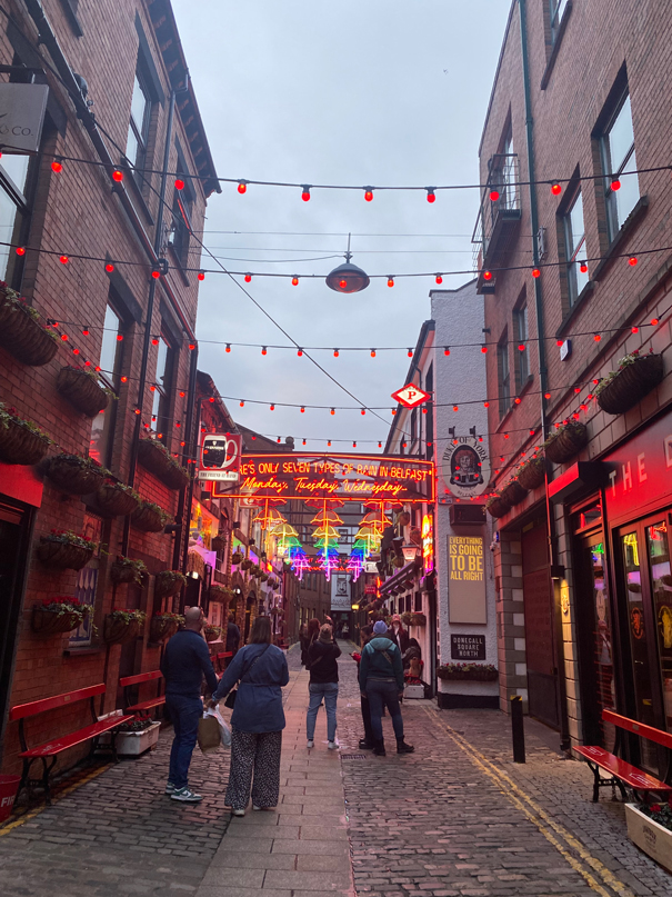 A lively cobblestone street in Belfast with illuminated signs, multiple persons, and red lights strung overhead, creating a vibrant and energetic atmosphere.