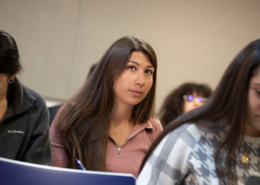Three people sitting, focused and writing, in a classroom setting. One person is looking up thoughtfully, holding a pen.