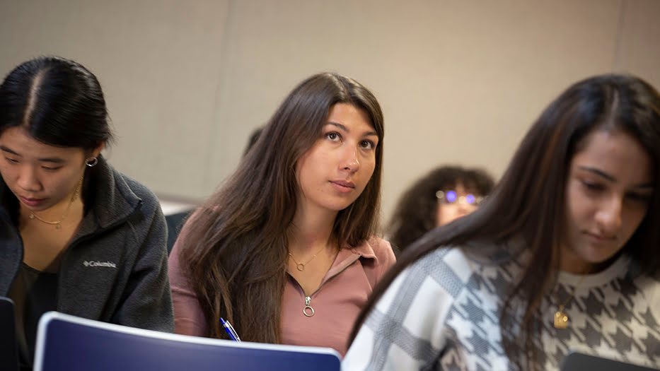 Three people sitting, focused and writing, in a classroom setting. One person is looking up thoughtfully, holding a pen.