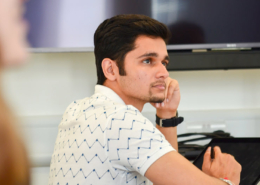 A person in a white, patterned shirt is attentively sitting at a table, looking towards a screen in a modern setting.
