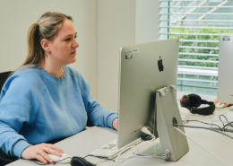 A person in a blue sweater works at a desk with an Apple computer, headphones nearby, and sunlight streaming through window blinds.