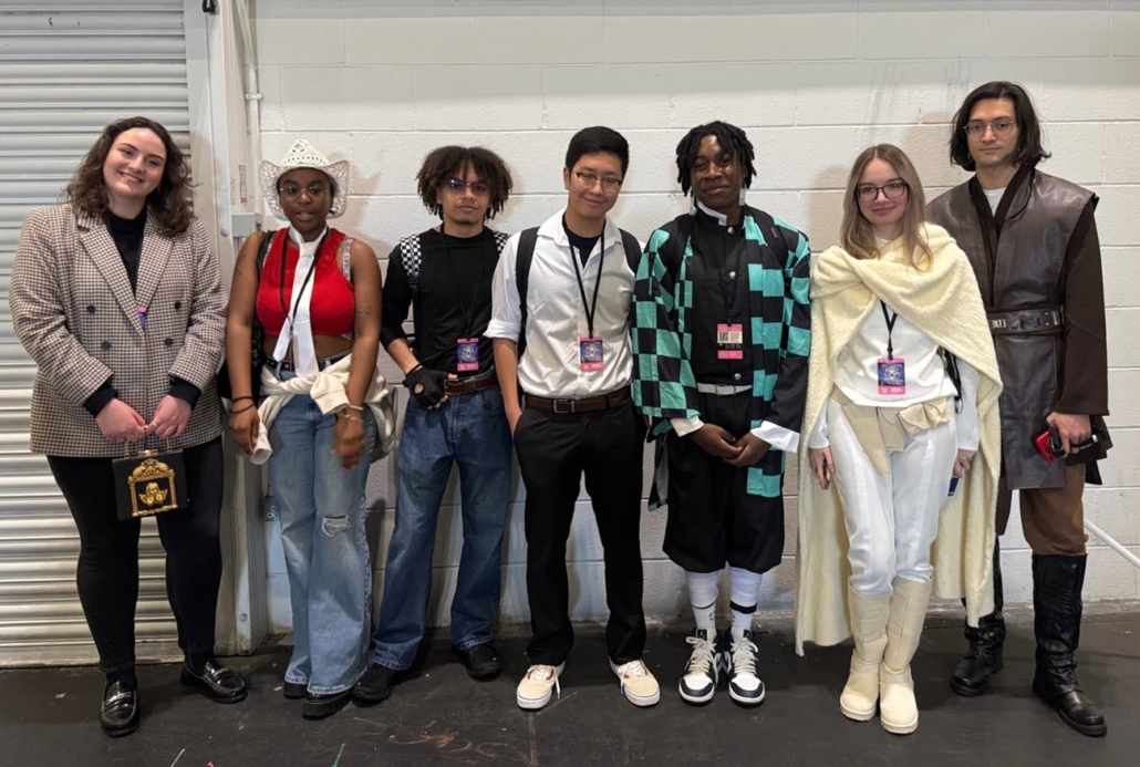 Seven cosplaying individuals stand side by side, posing indoors against a white wall, each displaying distinct attire and badges.