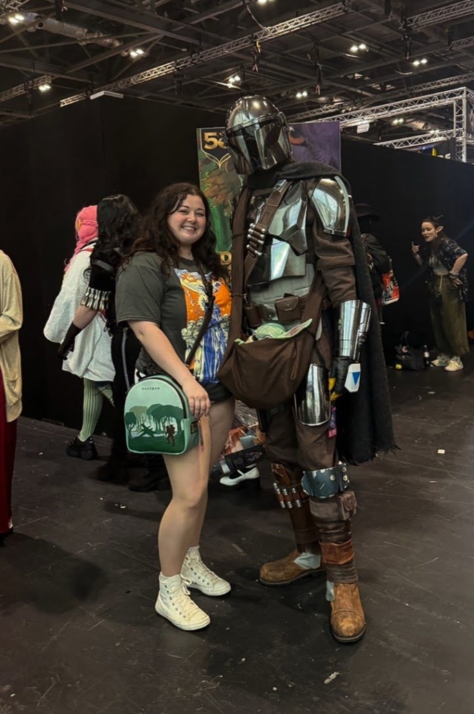A person poses with a costumed character at a convention, surrounded by several others in cosplay, under a well-lit event hall setting.