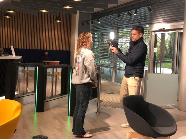 A person operates a camera with a flash, facing another person indoors. Modern interior, seating, and entryway visible in the background.