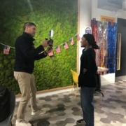 Two people interact in a room with a green wall, colorful decorations, and American flags. A person stands behind them near a doorway.