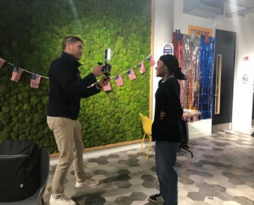 Two people interact in a room with a green wall, colorful decorations, and American flags. A person stands behind them near a doorway.