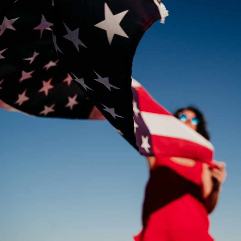 A person in sunglasses holds an American flag against a clear blue sky, wearing a red outfit. The flag is prominently displayed.