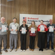 Six people are holding certificates in front of a Richmond American University London sign, standing indoors against a wooden backdrop.