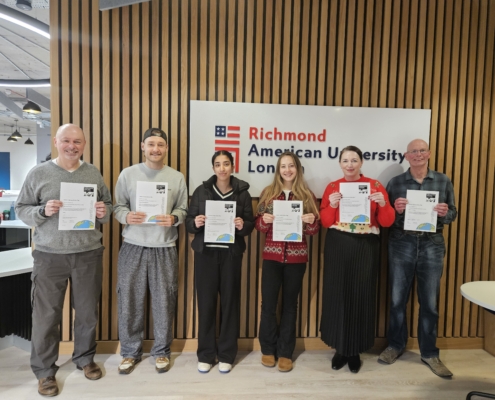 Six people are holding certificates in front of a Richmond American University London sign, standing indoors against a wooden backdrop.