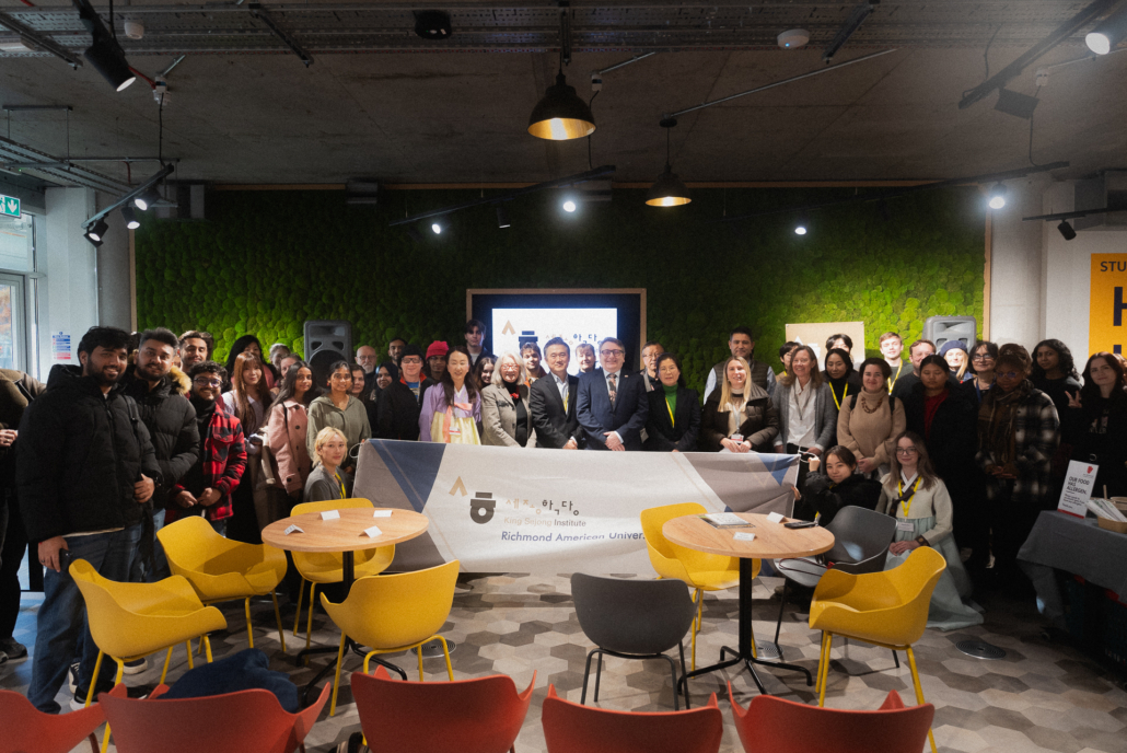 A group of people pose indoors holding a King Sejong Institute banner. The setting includes colorful chairs and a textured green wall.