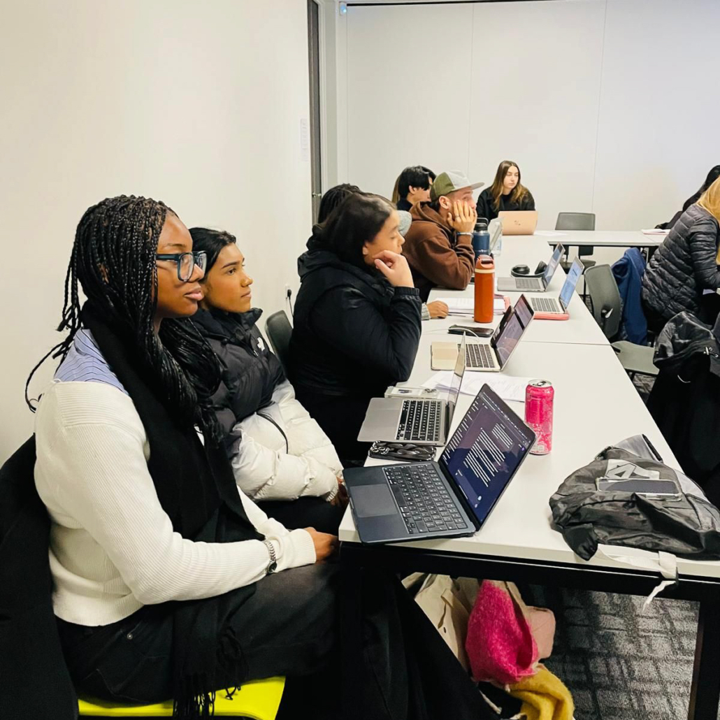 A group of people attentively listening in a classroom setting, each with a laptop on a white table, chairs arranged neatly.
