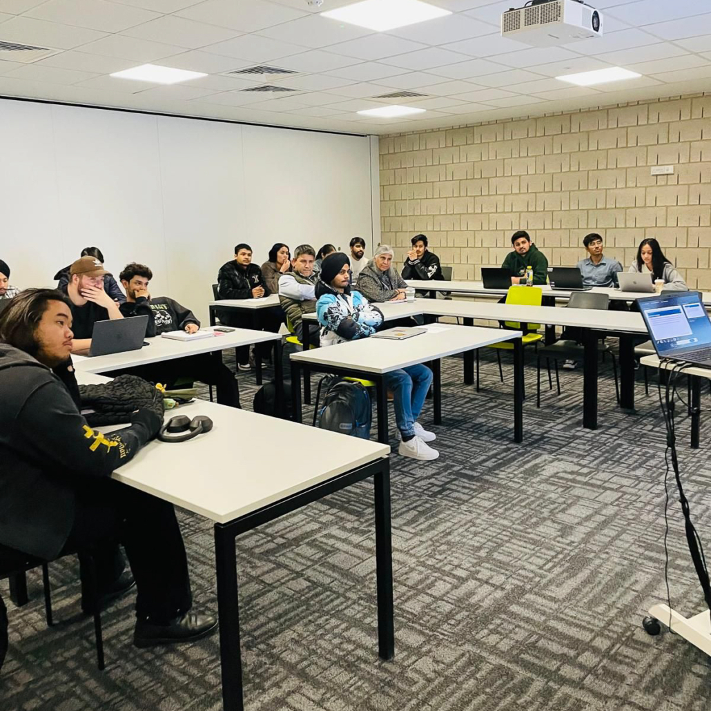 A classroom with people seated at desks, using laptops and paying attention to a presentation on a projector screen.