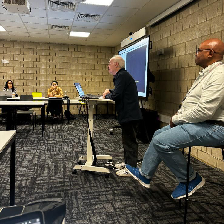 A person presents at a stand-up desk, engaging an audience of three seated individuals in a classroom setting with projector and screen.