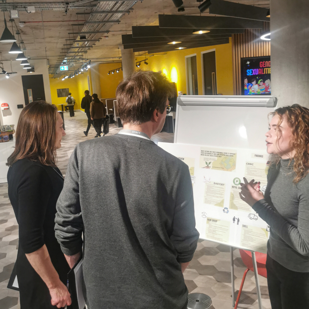 Three people in a modern, brightly lit office discuss charts on a whiteboard. Background features vibrant yellow walls and geometric flooring.
