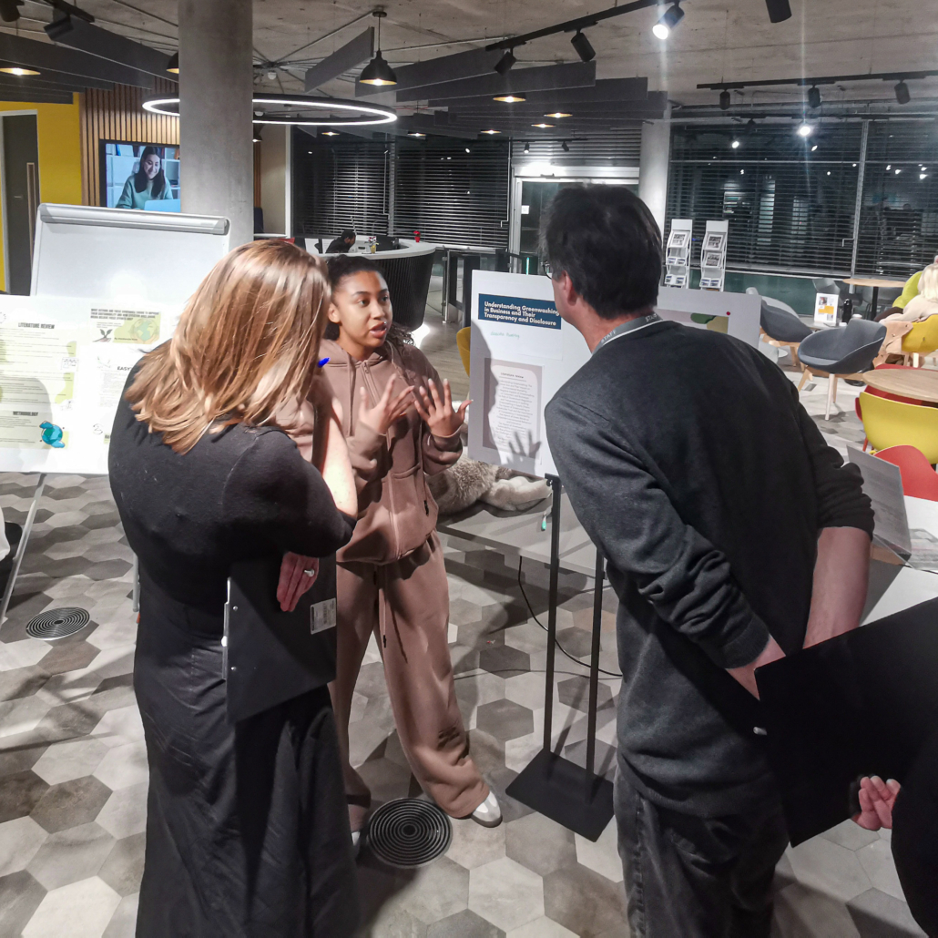 A person presents research on a board to two others in a modern, well-lit office with hexagonal floor tiles and presentation displays.