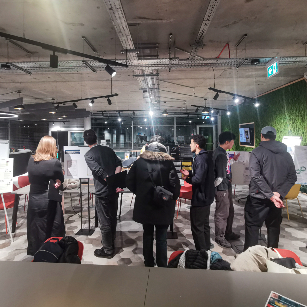 A group of people examines various displays and posters in a modern, industrial-style room with overhead lighting and a green feature wall.