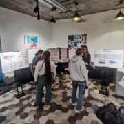 People are gathered in a room, discussing information displayed on posters. The setting has a modern design with hexagonal floor patterns.