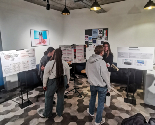 People are gathered in a room, discussing information displayed on posters. The setting has a modern design with hexagonal floor patterns.