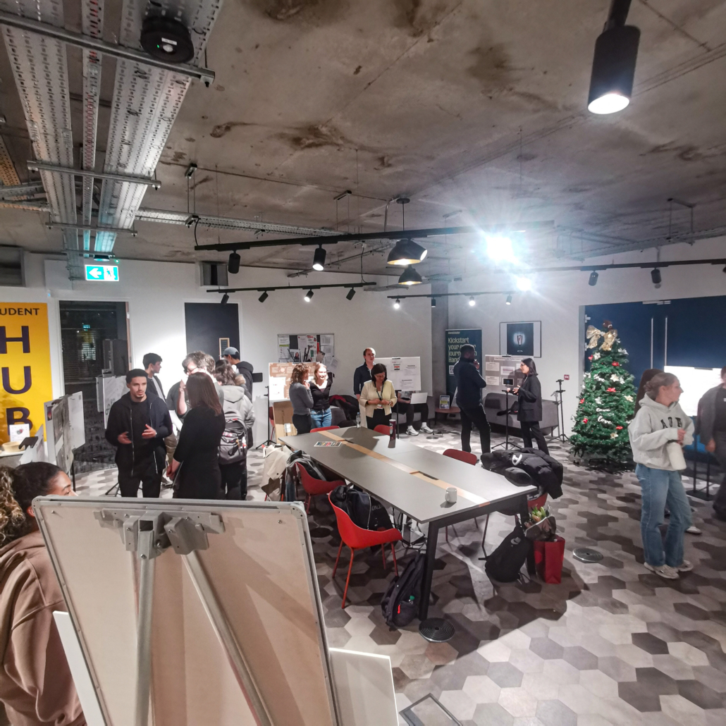 Indoor gathering with people conversing around tables. Posters on walls. Christmas tree in corner. Modern interior with exposed ceiling and geometric flooring.