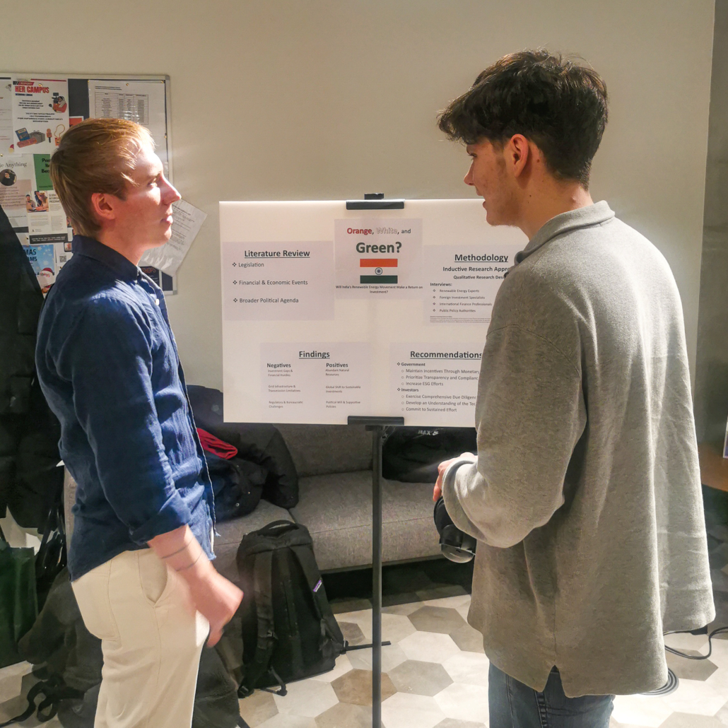 Two people are discussing a presentation board with research findings and recommendations, indoors, near a bulletin board, beside backpacks on a couch.